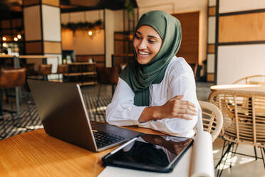 Muslim university student smiling happily while having a video conference with her tutor in a cafe. Cheerful woman with a hijab attending an online class in a coffee shop. - JLPSF25103