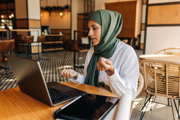 Young Muslim businesswoman having a discussion with her clients on a video call. Female entrepreneur with a hijab having an online meeting while working in a cafe. - JLPSF25104