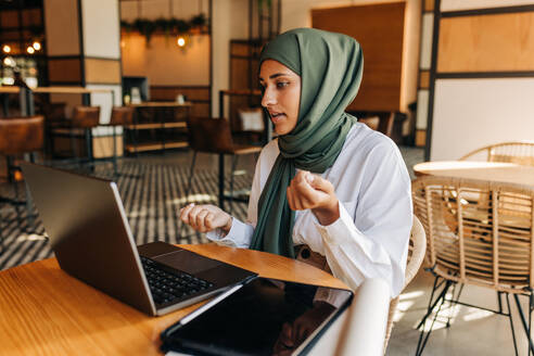 Junge muslimische Geschäftsfrau, die sich per Videoanruf mit ihren Kunden unterhält. Unternehmerin mit Hijab, die in einem Café eine Online-Sitzung abhält. - JLPSF25104