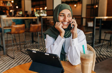 Cheerful Muslim businesswoman taking a phone call while sitting in a cafe with a digital tablet. Happy woman with a hijab speaking to her clients while working remotely. - JLPSF25106