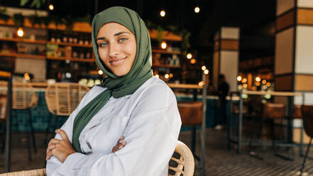 Portrait of a beautiful Muslim woman looking at the camera while sitting in a cafe. Young woman with a hijab sitting in a coffee shop with her arms crossed. - JLPSF25107