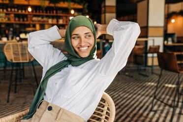 Nachdenkliche muslimische Frau, die mit einem glücklichen Lächeln wegschaut, während sie in einem Café sitzt. Fröhliche Frau mit Hijab, die nachdenklich ist, während sie sich in einem Café entspannt. - JLPSF25108