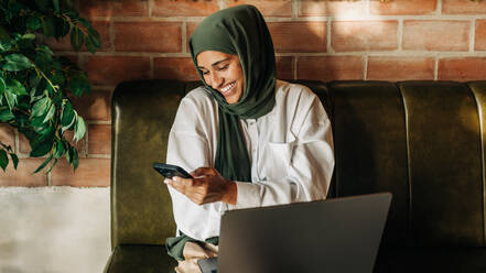 Businesswoman with a hijab smiling happily while using a smartphone in a cafe. Cheerful Muslim woman reading a text message while sitting on a couch with a laptop. - JLPSF25110