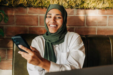 Young businesswoman with a hijab smiling at the camera cheerfully while holding a smartphone. Happy young Muslim woman doing remote work in a cafe. - JLPSF25111