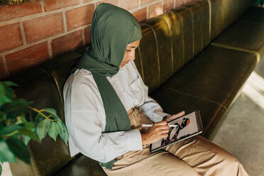 Muslimische Grafikdesignerin, die in einem Café ein kreatives Bild auf einem digitalen Tablet zeichnet. Junge Frau mit Hidschab, die auf einer Couch sitzend an einem neuen freiberuflichen Projekt arbeitet. - JLPSF25112