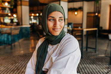 Nachdenkliche muslimische Frau, die wegschaut, während sie allein in einem Café sitzt. Nachdenkliche Frau mit einem Hijab, die sich in einem Café entspannt. - JLPSF25115