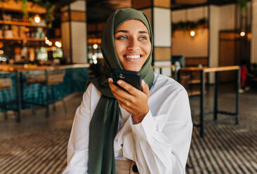 Lächelnde muslimische Geschäftsfrau beim Telefonieren in einem Café. Glückliche Frau mit Hijab, die mit ihren Kunden kommuniziert, während sie aus der Ferne arbeitet. - JLPSF25118