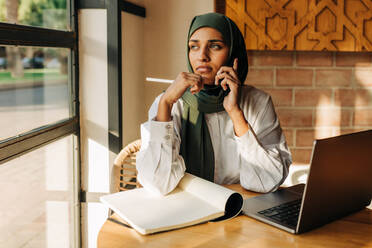 Studentin mit Hijab, die in einem Café telefoniert, während sie lernt. Konzentrierte muslimische Frau, die sich in einem Café auf dem Campus auf eine Prüfung vorbereitet. - JLPSF25121