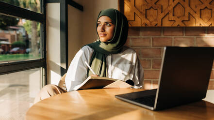 Muslim university student looking away thoughtfully while writing in a book. Young woman with a hijab preparing for an exam while sitting in a campus cafe. - JLPSF25122