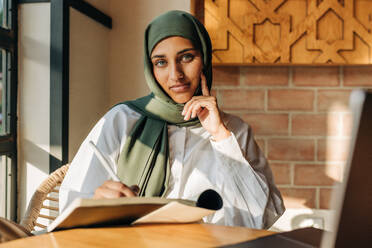 Female university student with a headscarf looking at the camera while writing in a book. Young Muslim woman preparing for an exam while sitting in a campus cafe. - JLPSF25123