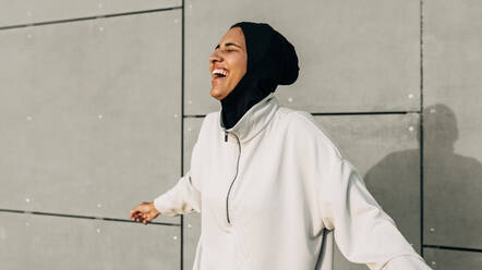 Sportliche muslimische Frau, die fröhlich lacht, während sie im Freien an einer Betonwand steht. Sportliche Frau mit Hijab, die während ihres morgendlichen Workouts Spaß hat. - JLPSF25133