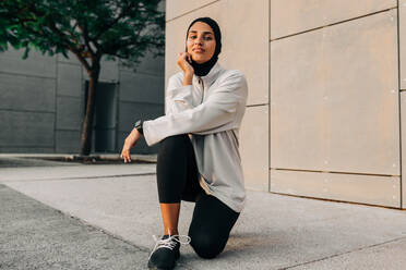 Sportliche Frau mit Hidschab, die in die Kamera schaut, während sie auf einem Betonboden im Freien kniet. Sportliche muslimische Frau beim morgendlichen Lauf in der Stadt. - JLPSF25137