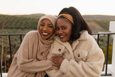 Glückliche beste Freundinnen, die im Freien in die Kamera lächeln. Zwei unterschiedliche junge Frauen, die sich gegenseitig umarmen, während sie sich an eine Brücke lehnen. Fröhliche Freundinnen, die am Wochenende zusammen abhängen. - JLPSF25148