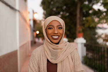 Glückliche junge muslimische Frau, die in die Kamera lächelt. Fröhliche muslimische Frau, die bei Sonnenuntergang auf einem Bürgersteig steht. Bescheidene junge Frau, die einen Hijab trägt. - JLPSF25153