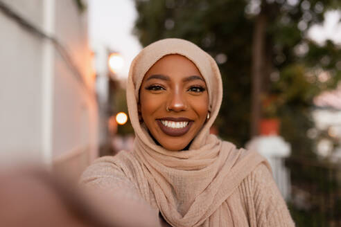 Fröhliche muslimische Frau, die lächelt, während sie ein Selfie im Freien macht. Glückliche muslimische Frau, die bei Sonnenuntergang auf einem Bürgersteig steht. Bescheidene junge Frau, die einen Hijab trägt. - JLPSF25154