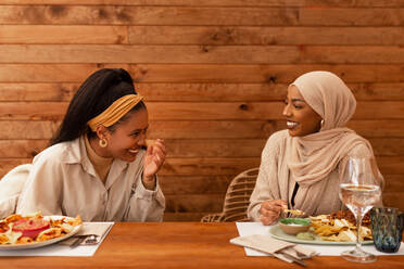 Beste Freundinnen lachen fröhlich beim Essen in einem Restaurant. Zwei unterschiedliche junge Frauen genießen ein gesundes Mittagessen in einer Cafeteria. Fröhliche Freundinnen, die am Wochenende zusammen abhängen. - JLPSF25165