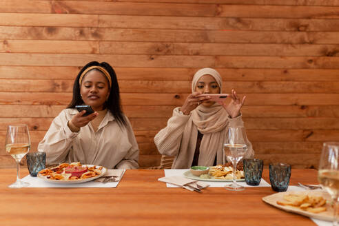 Zwei Freundinnen, die ihr Essen in einem Restaurant fotografieren. Zwei junge Frauen, die ein gesundes Mittagessen in einer Cafeteria einnehmen. Millennial-Freundinnen, die Inhalte für ihre Social-Media-Blogs erstellen. - JLPSF25166
