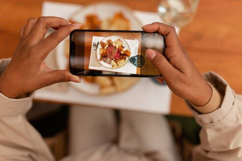 Bloggerin, die ihr Essen in einem Restaurant fotografiert. Hochformatige Ansicht einer nicht erkennbaren jungen Frau, die ein Fotohandy über ein Tablett mit Essen hält. Frau, die Inhalte für ihren Social Media Food Blog erstellt. - JLPSF25168