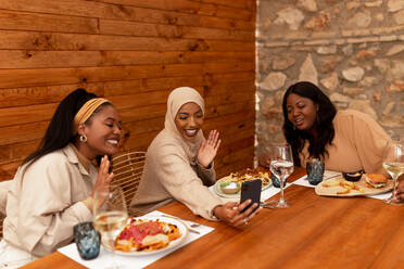 Ethnische Freundinnen winken sich während eines Videogesprächs zu. Glückliche junge Frauen lächeln, während sie mit ihren Freunden am Telefon sprechen. Gruppe multikultureller Freunde beim Mittagessen in einem Restaurant. - JLPSF25172