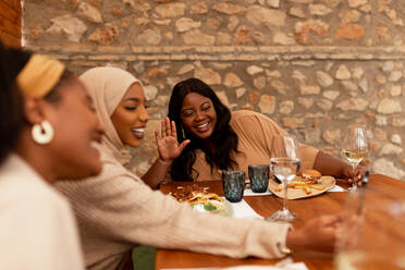 Fröhliche Freundinnen, die sich während eines Videogesprächs zuwinken. Fröhliche junge Frauen, die lächelnd mit ihren Freunden telefonieren. Gruppe multikultureller Freunde beim Mittagessen in einem Restaurant. - JLPSF25173