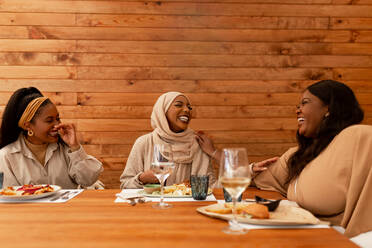 Fröhlich lachende Freunde bei einer Mahlzeit. Eine Gruppe glücklicher junger Frauen, die sich in einem Restaurant amüsieren. Drei multikulturelle Freunde bei einem gemütlichen Mittagessen am Wochenende. - JLPSF25178