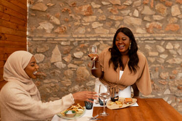 Ein Toast auf die Freundschaft: Zwei glückliche Freundinnen stoßen in einem Restaurant auf ihre Freundschaft an. Zwei unterschiedliche junge Frauen, die am Wochenende ein gemütliches Mittagessen genießen. - JLPSF25180