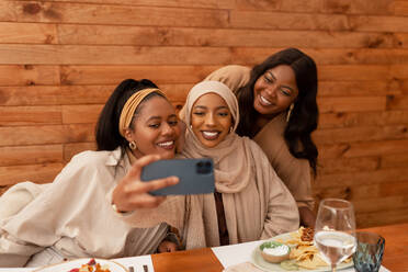 Multikulturelle Freundinnen machen ein gemeinsames Selfie in einem Restaurant. Glückliche junge Frauen lächeln fröhlich für ein Gruppenfoto. Verschiedene junge Freundinnen genießen ein Wiedersehensessen am Wochenende. - JLPSF25187