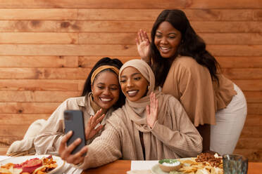 Millennial-Freundinnen winken sich während eines Videoanrufs zu. Glückliche junge Frauen lächeln, während sie mit ihren Freunden am Telefon sprechen. Gruppe multikultureller Freundinnen beim Mittagessen in einem Restaurant. - JLPSF25189