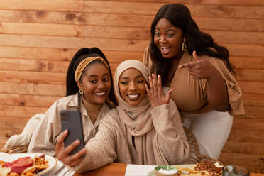 Frisch verlobte muslimische Frau zeigt ihren Ring bei einem Videoanruf. Gruppe von drei multikulturellen jungen Frauen, die ihre Verlobung in einem Restaurant feiern. Fröhliche Freundinnen beim gemeinsamen Mittagessen. - JLPSF25190