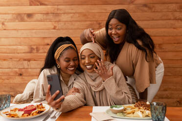 Verlobte muslimische Frau zeigt ihren Ring bei einem Videoanruf. Gruppe von drei multikulturellen jungen Frauen, die in einem Restaurant ihre Verlobung feiern. Fröhliche Freundinnen beim gemeinsamen Mittagessen. - JLPSF25191