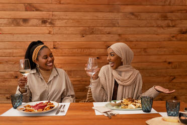 Junge Freundinnen genießen ein feierliches Mittagessen in einem Restaurant. Zwei glückliche Freundinnen lächeln und halten ihre Trinkgläser hoch. Zwei unterschiedliche junge Frauen essen am Wochenende zusammen. - JLPSF25192