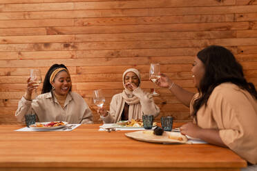 Ich bringe einen Toast aus. Drei fröhliche Freundinnen, die mit Gläsern in einem Restaurant feiern. Drei multikulturelle junge Frauen, die ein gemütliches Mittagessen am Wochenende genießen. - JLPSF25193