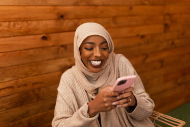 Excited muslim woman sending a text message while sitting in a restaurant. Happy young muslim woman smiling cheerfully while using her smartphone. Carefree young woman having fun in a cafe. - JLPSF25200