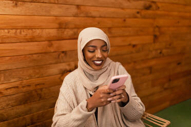 Cheerful muslim woman sending a text message while sitting in a restaurant. Happy young muslim woman smiling excitedly while using her smartphone. Carefree young woman having fun in a cafe. - JLPSF25201