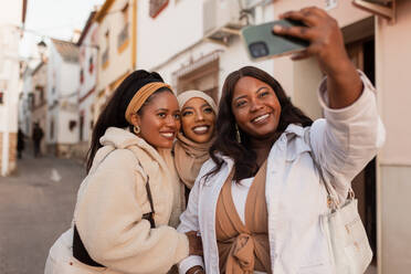 Drei multikulturelle Freundinnen machen ein Selfie im Freien. Glückliche junge Frauen, die für ein Gruppenfoto lächeln, während sie zusammen stehen. Eine Gruppe von verschiedenen Freunden, die ein Wiedersehen am Wochenende genießen. - JLPSF25211