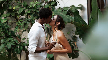Leidenschaftliches junges Paar, das sich küsst, während es ein Glas Sekt in der Hand hält. Junges Paar, das einen romantischen Moment in seinem Garten teilt. Interrassisches Paar, das seinen Jahrestag zu Hause feiert. - JLPSF25231
