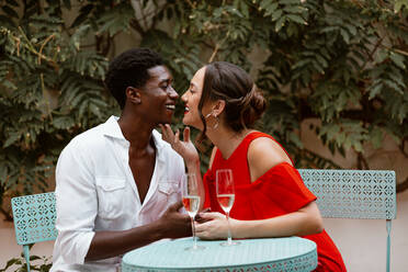 Lächelndes Paar, das sich im Hinterhof eines Ferienhauses küsst. Romantisches Paar, das seinen Jahrestag mit Sekt feiert. Fröhliches junges Paar, das sich an einem Tisch im Freien entspannt. - JLPSF25237
