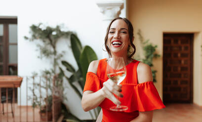 Cheerful young woman toasting with a wineglass on a balcony. Happy young woman celebrating while standing outside a holiday home. Woman laughing and having a good time on the weekend. - JLPSF25252