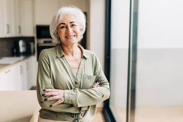 Elderly woman smiling happily while standing indoors. Cheerful senior woman enjoying her retirement years in her home. Senior woman looking at the camera with her arms crossed. - JLPSF25275