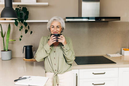 Ältere Frau genießt eine Tasse Kaffee, während sie ihren Tag in ihrem Tagebuch plant. Sorglose ältere Frau schaut in die Kamera, während sie in ihrer Küche zu Hause steht. - JLPSF25279
