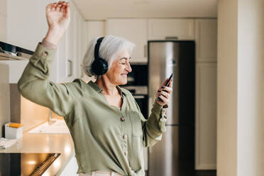 Fröhliche ältere Frau, die zu ihrer Lieblingsmusik tanzt, während sie drahtlose Kopfhörer trägt. Fröhliche ältere Frau, die eine gute Zeit hat, während sie Musik auf ihrem Smartphone spielt. - JLPSF25282