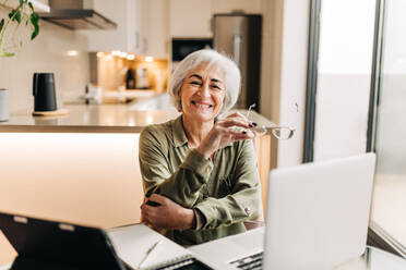 Reife Geschäftsfrau, die in die Kamera lächelt, während sie an ihrem Schreibtisch in ihrem Heimbüro sitzt. Erfolgreiche ältere Geschäftsfrau, die die Arbeit von zu Hause aus genießt. - JLPSF25284