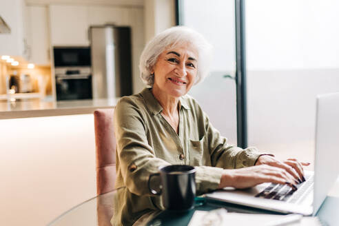 Grauhaarige ältere Geschäftsfrau, die an ihrem Schreibtisch sitzend auf ihrem Laptop tippt. Erfolgreiche Geschäftsfrau, die fröhlich lächelt und die Arbeit von zu Hause aus genießt. - JLPSF25286