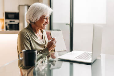 Glückliche ältere Geschäftsfrau, die sich während einer Videokonferenz mit ihren Geschäftspartnern Notizen macht. Reife Geschäftsfrau, die glücklich lächelt, während sie an einer Sitzung in ihrem Heimbüro teilnimmt. - JLPSF25287