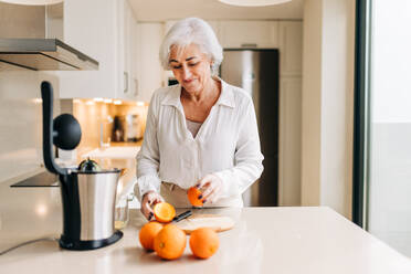 Fröhliche ältere Frau, die in ihrer Küche zu Hause mit einem elektrischen Mixer Fruchtsaft herstellt. Fröhliche ältere Frau, die sich im Ruhestand gesund und nahrhaft ernährt. - JLPSF25297