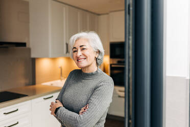 Silver-haired woman smiling at the camera while standing with her arms crossed. Cheerful senior woman enjoying her retirement years in her home. - JLPSF25302