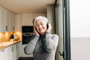 Ältere Frau, die ihre Lieblingsmusik über kabellose Kopfhörer hört. Glückliche ältere Frau, die Spaß hat und ihren Ruhestand zu Hause genießt. - JLPSF25305