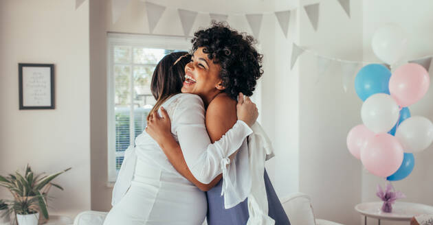 Pregnant woman hugging her best female friend at baby shower. Pregnant woman celebrating baby shower at home with best friends. - JLPSF25311