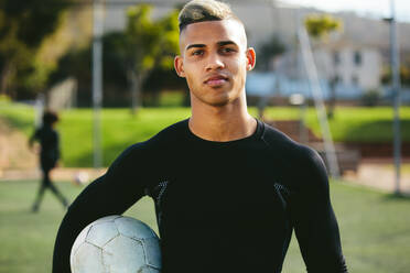 Porträt eines jugendlichen Fußballspielers, der einen Fußball in der Hand hält und in die Kamera schaut. Junger Mann beim Training auf dem Fußballplatz. - JLPSF25319