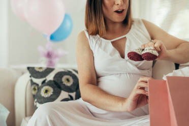 Schwangere Frau sitzt auf dem Sofa und öffnet Geschenke bei einer Babyparty. Frau hält neue Babyschühchen zu Hause. - JLPSF25342